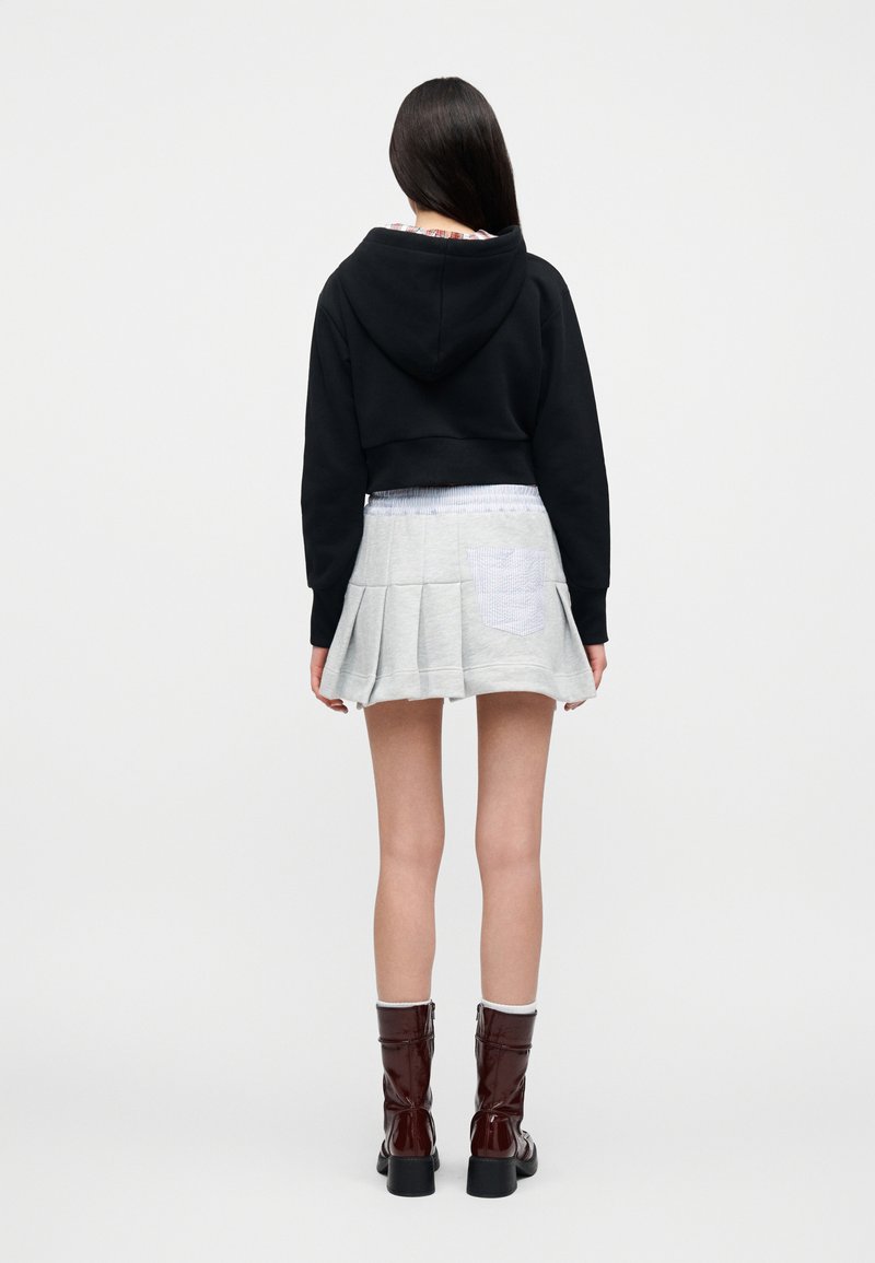 Sweat à capuche noir coupé, jupe plissée grise avec une poche à carreaux, bottines marron brillantes à talons blocs, et chaussettes côtelées.