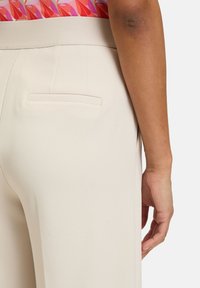 Beige skräddarsydda byxor med en slät textur, som har en diskret bakficka och ett brett midjeband. Byxorna har en figurnära silhuett.