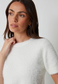 Kobieta z brązowymi włosami do ramion, ubrana w białą, puszystą bluzkę z krótkim rękawem, patrzy lekko w lewo z ręką przy szyi.