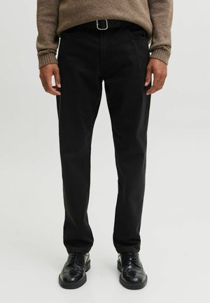 Homme portant un pantalon noir, des chaussures habillées en cuir noir et un pull marron, debout devant un fond clair uni.