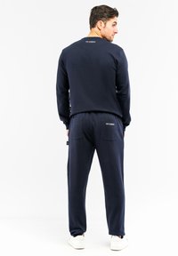 Mørkeblå sweatshirt- og bukser sæt med en afslappet pasform, blødt stof, ribbede manchetter og logo detalje på ryggen. Båret med hvide sneakers.