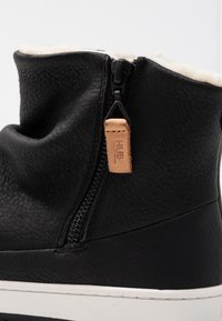 Bota de cuero negro con acabado texturizado, forro de felpa blanca, cierre de cremallera y etiqueta de acento de cuero marrón. Suela blanca resistente.