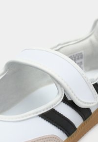 White and black athletic shoe with a textured tan sole, featuring a broad Velcro strap and contrasting stripes across the upper.