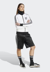 Witte sportieve zipjack met zwarte accenten, gecombineerd met zwarte shorts. Heeft drie strepen op de mouwen en zijkanten, en zwarte sneakers.