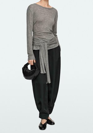 Osoba v dlouhém šedém topu s detaily vázání v pase, volných černých kalhotách, černých balerínách a s malou černou kabelkou.