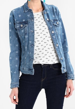 Kvinde iført en blå denimjakke med hjerte-mønstre, en hvid skjorte med små sorte cykler og mørkeblå jeans.