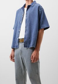 Camisa azul de mezclilla de manga corta con cuello, con botones delanteros. Combinada con pantalones de rayas verticales, que muestran un diseño informal.