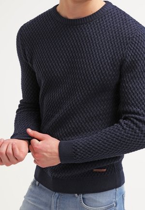 Persona che indossa un maglione di maglia scuro e strutturato, aggiustando la manica, con jeans azzurri visibili in vita.
