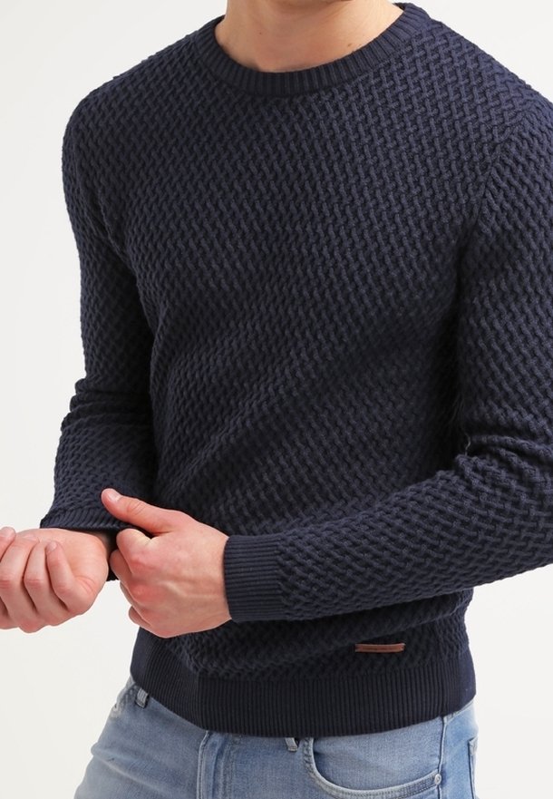 Personne portant un pull en tricot sombre texturé ajustant la manche, avec un jean bleu clair visible à la taille.