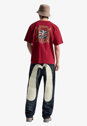Uomo che indossa una T-shirt rossa con grafica, jeans scuri con un grande motivo bianco curvo sul retro, cappello beige a secchiello e sneakers, in piedi di spalle.