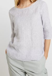 Žena v světlé šedé texturované blůze s tříčtvrtečními rukávy a světlých šedých kalhotách, s rukama v kapsách na jednobarevném pozadí.