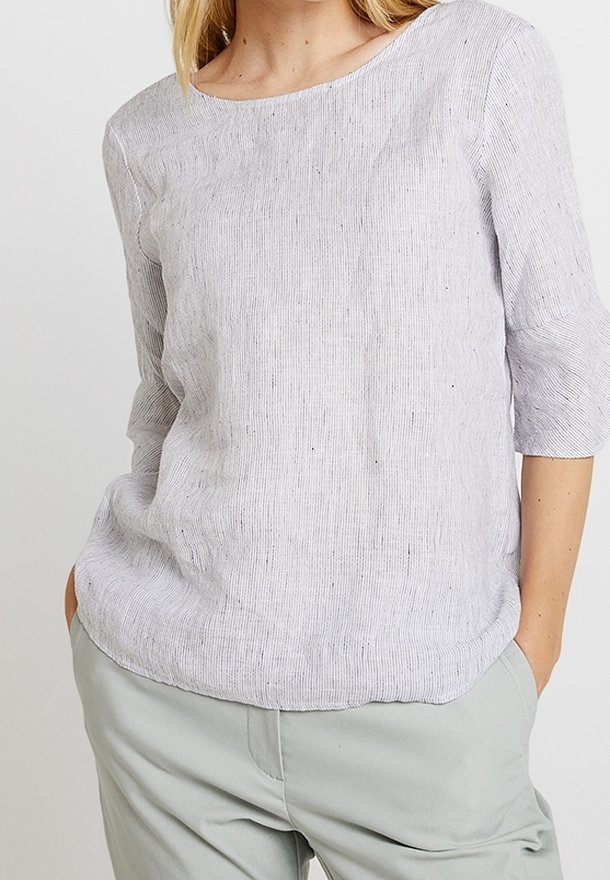 Žena v světlé šedé texturované blůze s tříčtvrtečními rukávy a světlých šedých kalhotách, s rukama v kapsách na jednobarevném pozadí.