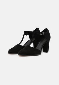 Escarpins en daim noir avec un design à bride en T, un bout pointu et un talon bloc. Accentué par une petite boucle en argent sur la bride.