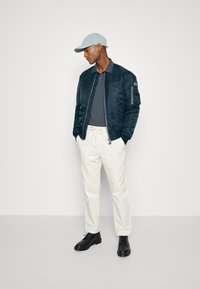 Giacca bomber navy con zip, polo grigia, pantaloni bianchi e scarpe nere. Cappello azzurro chiaro con visiera curva. Stile semplice e moderno.