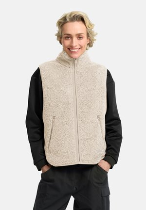 Lachend persoon met kort blond haar draagt een beige fleecevest over een zwart shirt met lange mouwen en een zwarte broek, handen in de zakken.