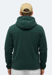 Felpa con cappuccio in felpa verde scuro, dalla superficie testurizzata, con cappuccio ampio e dettaglio delle cuciture sul retro, indossata con un cappello beige. Nessuna cerniera o accentuazione visibile.