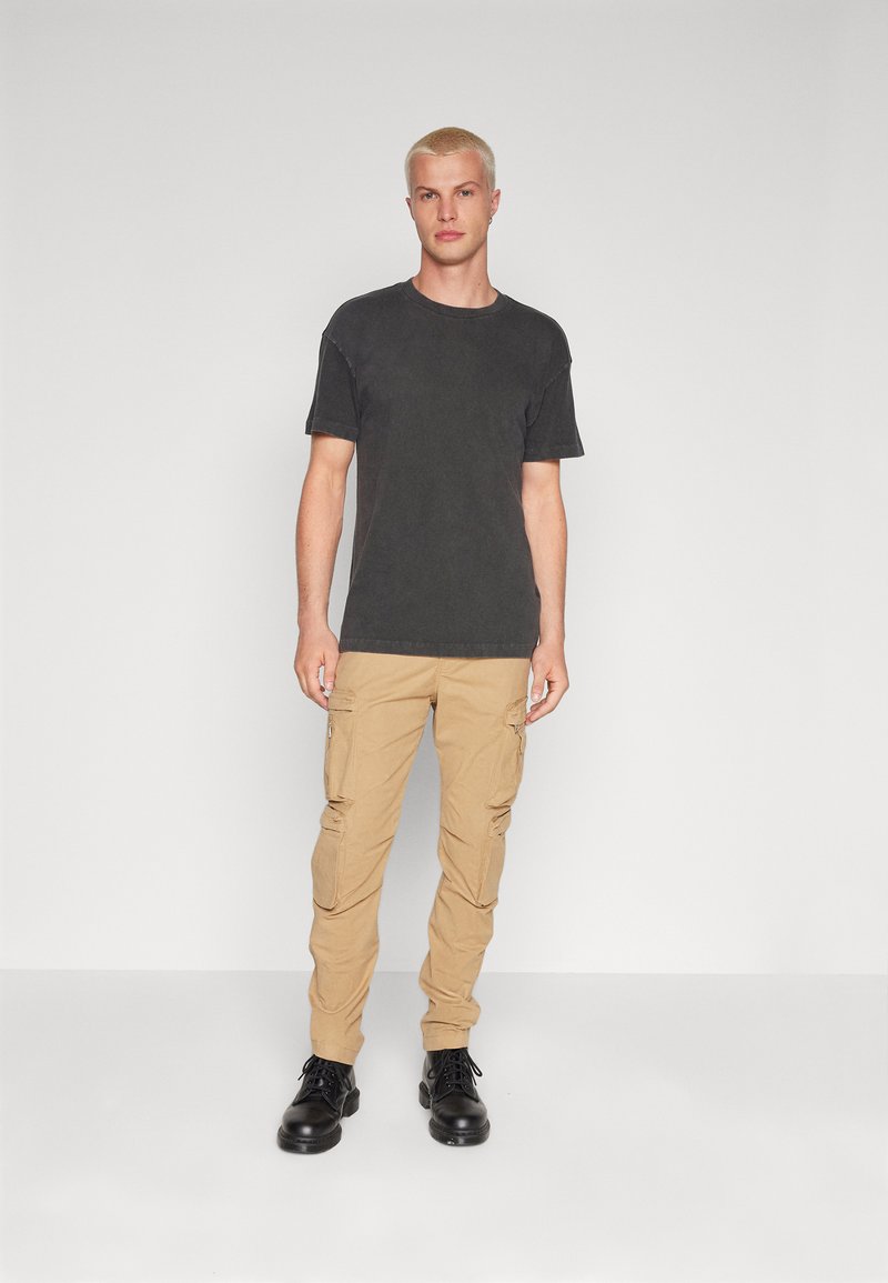 Junger Mann mit kurzen blonden Haaren, der ein schwarzes T-Shirt, beige Cargo-Hosen und schwarze Lederstiefel trägt, steht vor einem schlichten weißen Hintergrund.