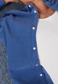 Hombre acostado sobre una alfombra azul, vistiendo una camisa azul abotonada sobre una camiseta sin mangas blanca acanalada.