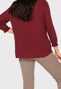 Burgundowa bluzka z rękawami 3/4, luźny krój i rozcięcie z tyłu, w połączeniu z jasnobrązowymi spodniami w prążki.