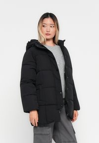 Joven mujer vestida con una chaqueta acolchada negra, suéter gris y pantalones cargo grises, mirando hacia su izquierda contra un fondo blanco liso.