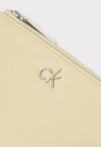 Pochette en faux cuir jaune avec un logo 'CK' en métal, dotée d'une fermeture éclair métallique et de couture texturée le long du bord.