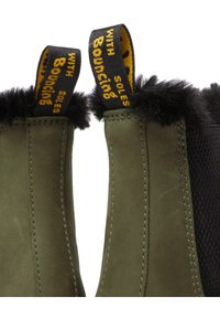 Close-up van olijfgroene laarzen met een zwarte bontvoering en zwarte aantreklussen met gele tekst "WITH BOUNCING SOLES".