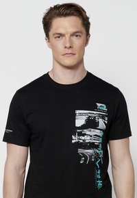 Juodas medvilnės marškinėlis su grafiniu dizainu, vaizduojančiu lenktynių automobilius monochrominėje paletėje su žydramis smulkiai. Apvalus kaklas ir trumpomis rankovėmis.