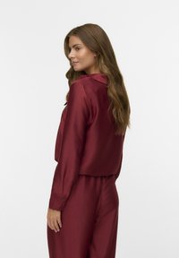 Burgundy, silky blouse with long sleeves, button closures, and a collared neckline; features a ribbed texture and relaxed fit.