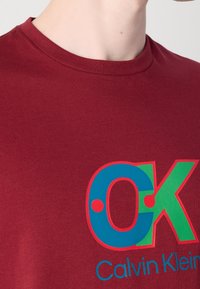 Gros plan d'une personne portant un t-shirt Calvin Klein bordeaux avec un grand graphisme multicolore "OK" sur la poitrine.