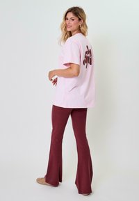 Růžové bavlněné tričko s krátkým rukávem a tmavě hnědou grafikou na zádech, doplněné vínovými zvonovými kalhotami a béžovou obuví.