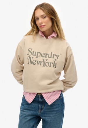 Felpa beige con la scritta "Superdry New York" in testo grigio scuro, indossata sopra una camicia a righe rosa e bianca, abbinata a jeans blu.