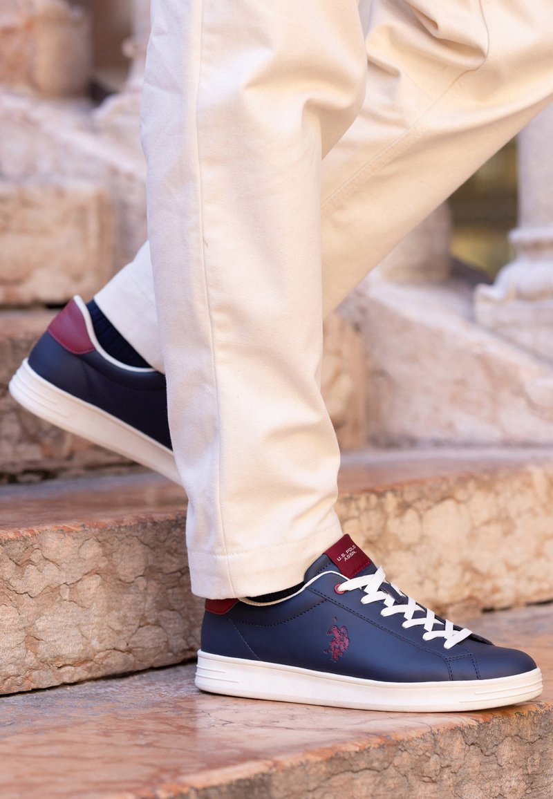Personne portant un pantalon blanc et des baskets bleues avec des lacets blancs marchant sur des escaliers en pierre à l'extérieur.