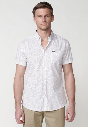 Camisa blanca de manga corta con un patrón texturizado de pequeños puntos negros, que presenta un cuello y un bolsillo en el pecho izquierdo con un logotipo.