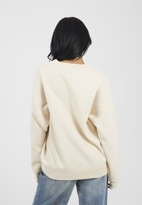 Persona con lunghi capelli neri che indossa una felpa color crema e jeans blu, rivolta di spalle contro uno sfondo bianco semplice.