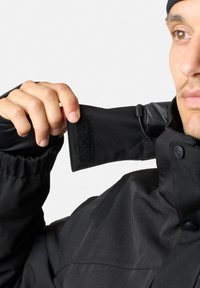 Zwarte jas met een hoge kraag en een klittenbandsluiting, knoopsluitingen en een gladde textuur. Mouwen zijn elastisch voor een goede pasvorm.