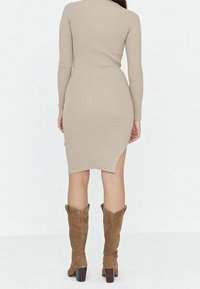 Robe beige à côtes avec manches longues, longueur médiane et fente sur le côté. Associée à des bottes en daim marron hauteur genoux avec des talons épais.