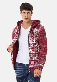 Bordeauxfarbene Zip-Up-Hoodie mit weißen Grafiken und Text, ausgestattet mit einer Zugkordelkapuze, gerippten Bündchen und einer einfarbigen weißen Innenschicht, kombiniert mit Jeans.