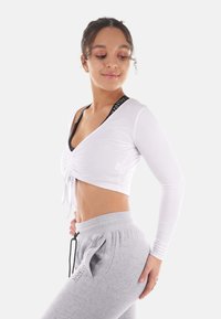 Weißes Langarm-Crop-Top mit gerafftem Frontdetail, kombiniert mit grauen Jogginghosen mit seitlichen Taschen und einem Kordelzugbund.