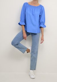 Blouse bleu clair à épaules dénudées avec un col smocké et des manches bouffantes, associée à un jean en denim clair et des baskets blanches.