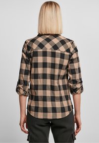 Chemise à carreaux en tissu beige et noir, à manches longues retroussées, présentant un ourlet courbé et un design de coupes au dos.