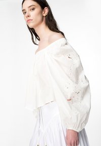 Top bianco in cotone a spalle scoperte con maniche a sbuffo. Presenta motivi floreali ricamati e dettagli in pizzo. Design largo e fluente.