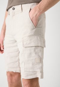 Beige linne cargo shorts med side lommer, rullet kant og et firkantet logo-patch. Har knappelukking og et avslappet design.