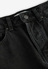 Gros plan sur la taille d'un jean en denim noir avec bouton, passants de ceinture et détails de poche avant sur fond blanc.