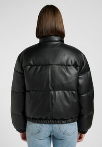 Mujer con el pelo liso castaño que lleva una chaqueta de cuero acolchada negra y jeans de color azul claro, de espaldas contra un fondo sencillo.