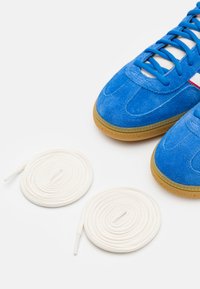 Modre semišnike s gumijastim podplati, belimi vezalkami in subtilnim rdečim poudarkom. Ob njih so še dve dodatni beli vezalki.