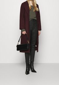 Bordowy wełniany płaszcz z szerokim kołnierzem, ciemnozielony sweter, czarne skórzane spodnie oraz czarne botki na szpilce, trzymając czarną torebkę.