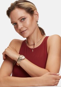 Guldtonet armbåndsur med en rund urskive, metalarmbånd og lyserød skive. To lagde sølvkæder med en spidst vedhæng. Rød ærmeløs top.