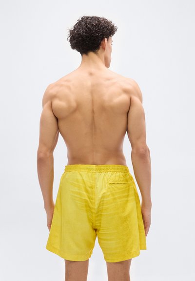 Άνδρας χωρίς πουκάμισο με σγουρά μαλλιά στέκεται με την πλάτη στραμμένη, φορώντας χαλαρά κίτρινα σορτς πάνω σε απλό λευκό φόντο.