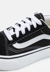 Zapatilla de lona negra con cordones blancos, detalles en forma de ola blanca y costuras negras. Presenta una suela de goma blanca con un acabado texturizado.