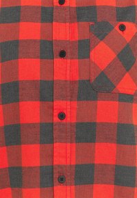 Red and gray plaid flannel shirt with black buttons, featuring a pocket, contrasting colors, and a textured fabric.